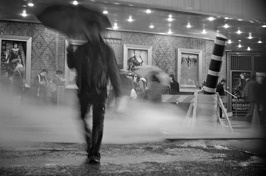 Black and white night street scene: a figure with an umbrella walks through steam past a theatre marquee in the rain.